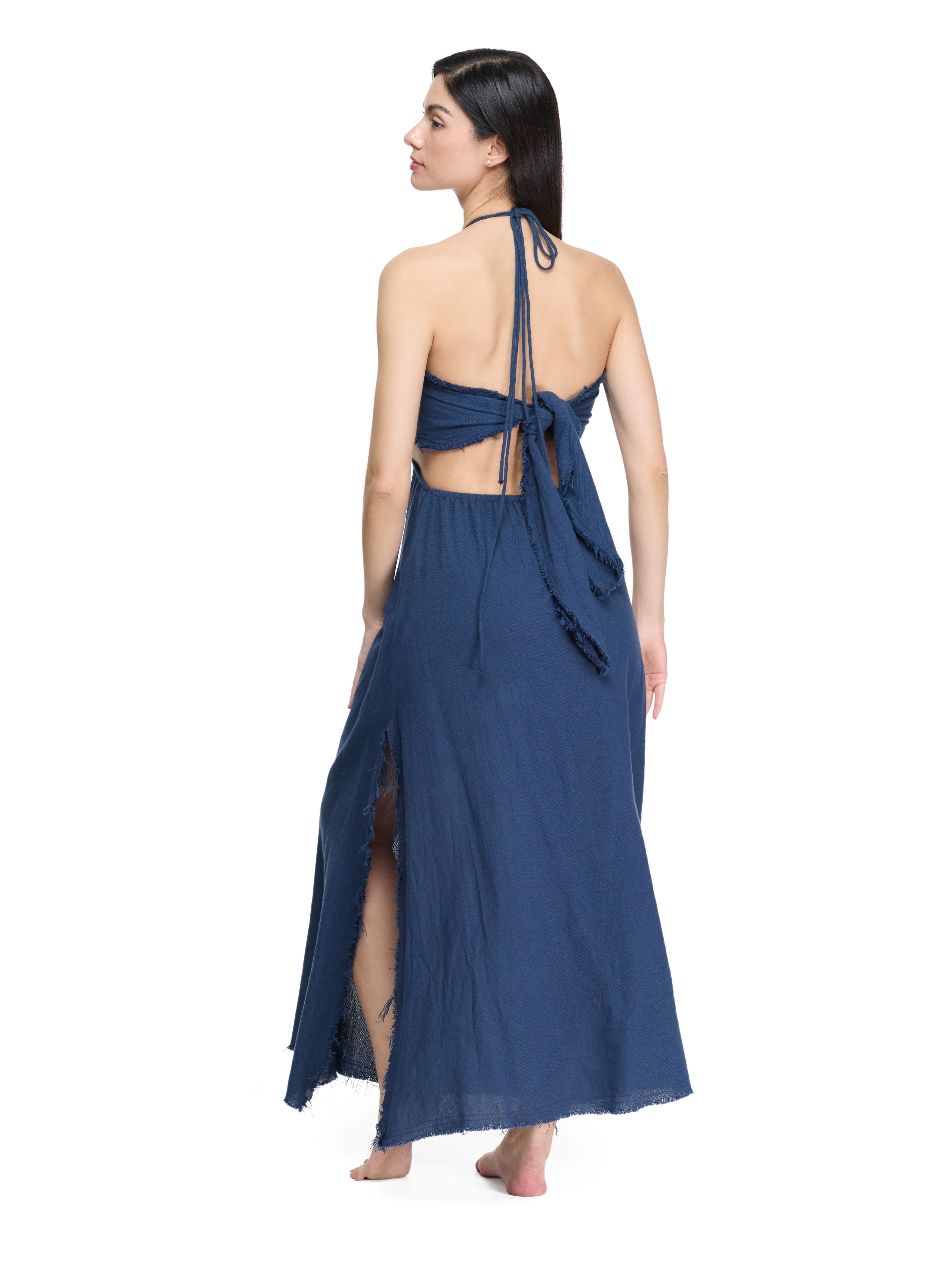 Back view of flowy navy maxi dress, open back with waist tie knot, halter cords, raw fringe hem, side slit skirt panel.