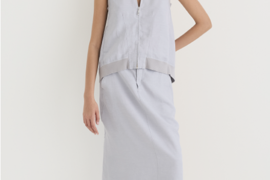 Sleeveless V-neck vest in misty gray linen with a zip front and contrast hem band, paired with a matching long asymmetric-hem skirt; styled with snakeskin-print mules against a