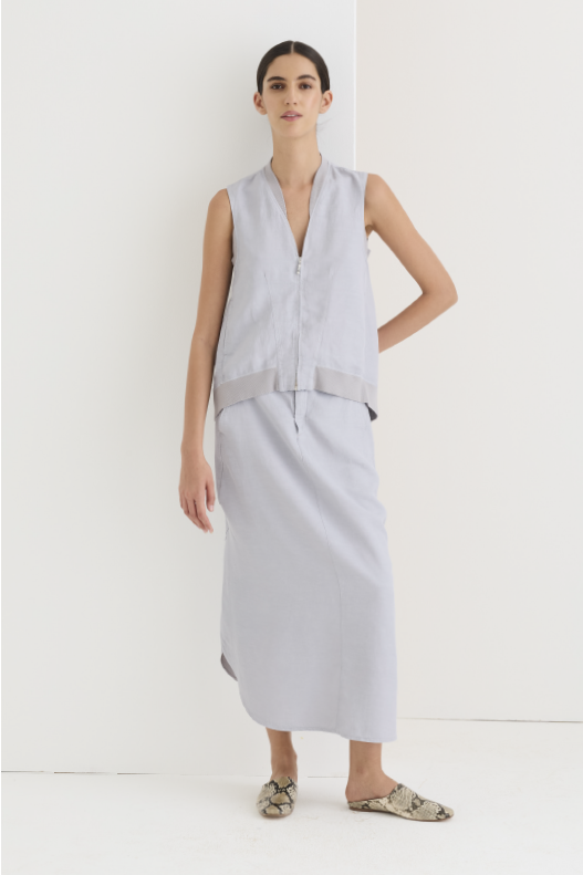 Sleeveless V-neck vest in misty gray linen with a zip front and contrast hem band, paired with a matching long asymmetric-hem skirt; styled with snakeskin-print mules against a