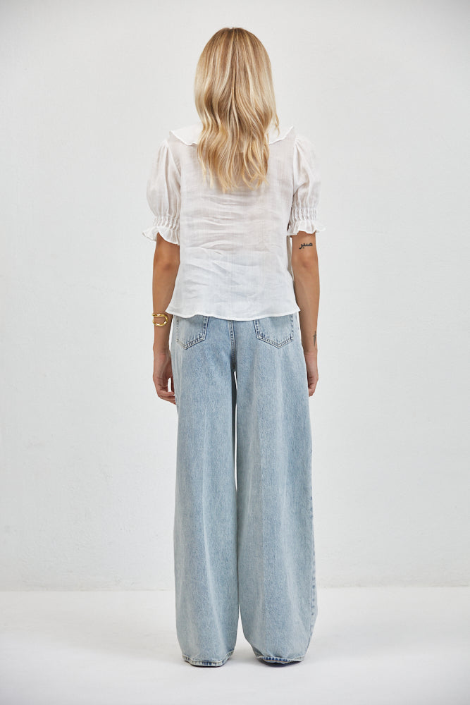Back view of sheer white cotton-linen top with smocked puff sleeves and ruffled hem, tucked into light-wash wide-leg jeans.