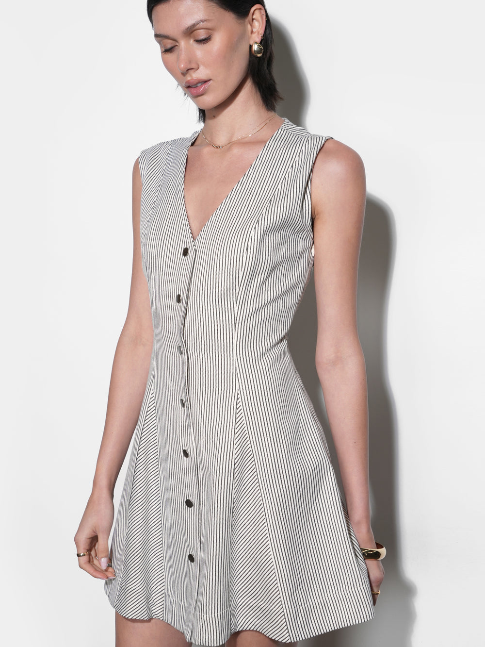 Sleeveless cream and black pinstripe mini dress with deep V-neckline, matte brass snap buttons, and angled panel skirt — fitted waist, front view, mid-thigh length.