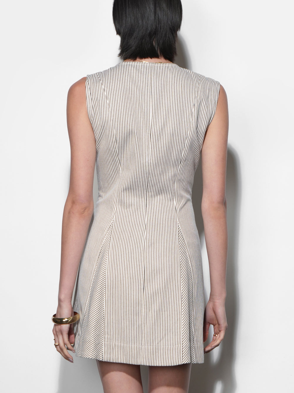 Back view of cream and near-black ticking stripe mini dress, sleeveless, structured canvas fabric, angled skirt panels, fitted waist, gold bangle bracelet detail.