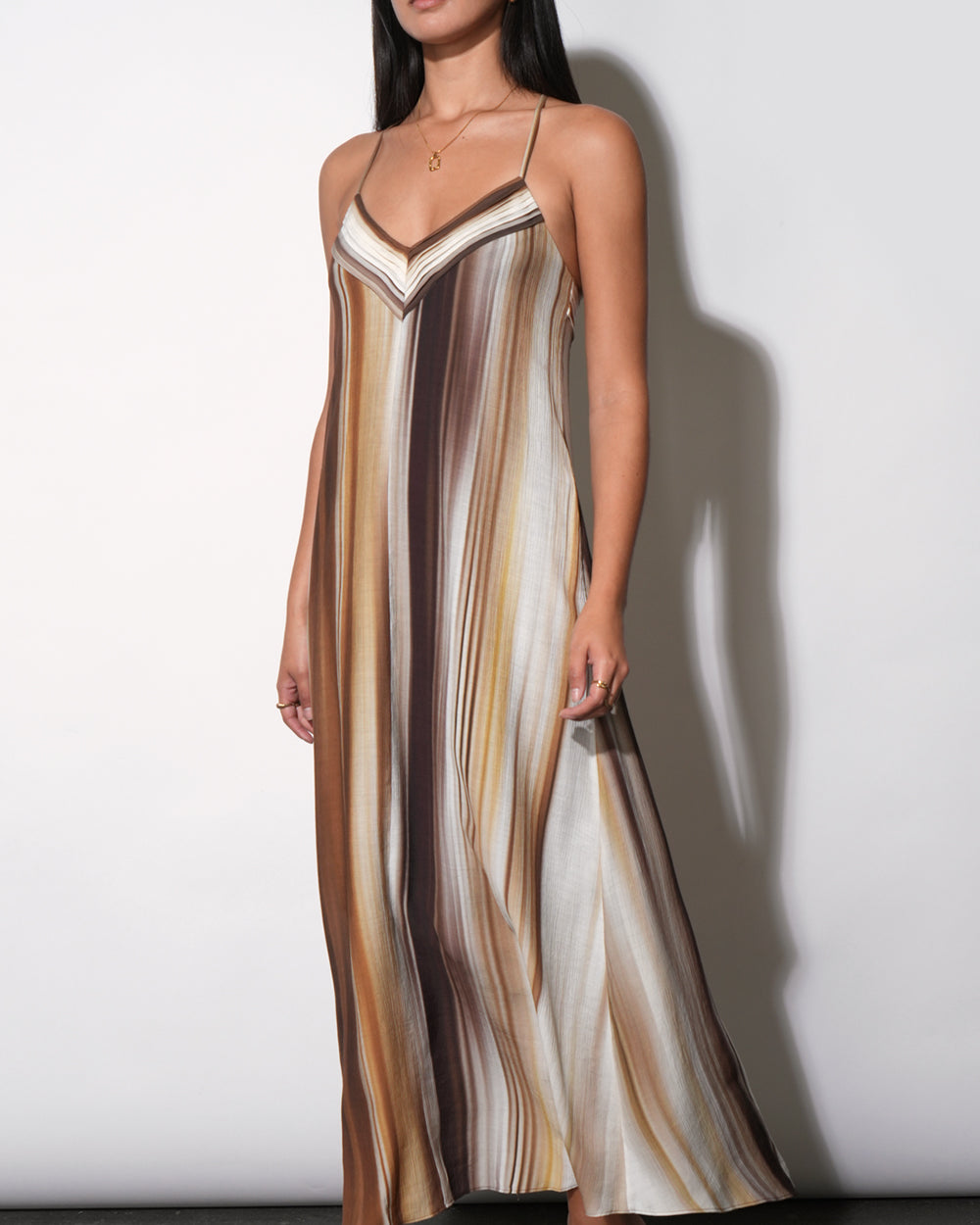 Fluid satin-finish maxi slip dress with blended vertical watercolor stripe print, V-neckline with contrast binding, spaghetti straps, full-length A-line skirt. Side profile. Styled with strappy
