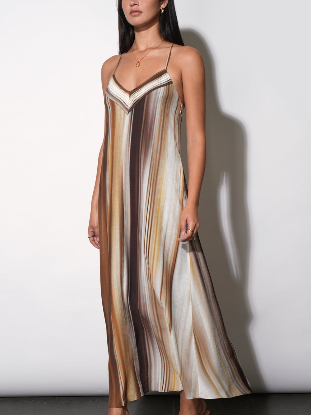 Fluid satin-finish maxi slip dress with blended vertical watercolor stripe print, V-neckline with contrast binding, spaghetti straps, full-length A-line skirt. Side profile. Styled with strappy