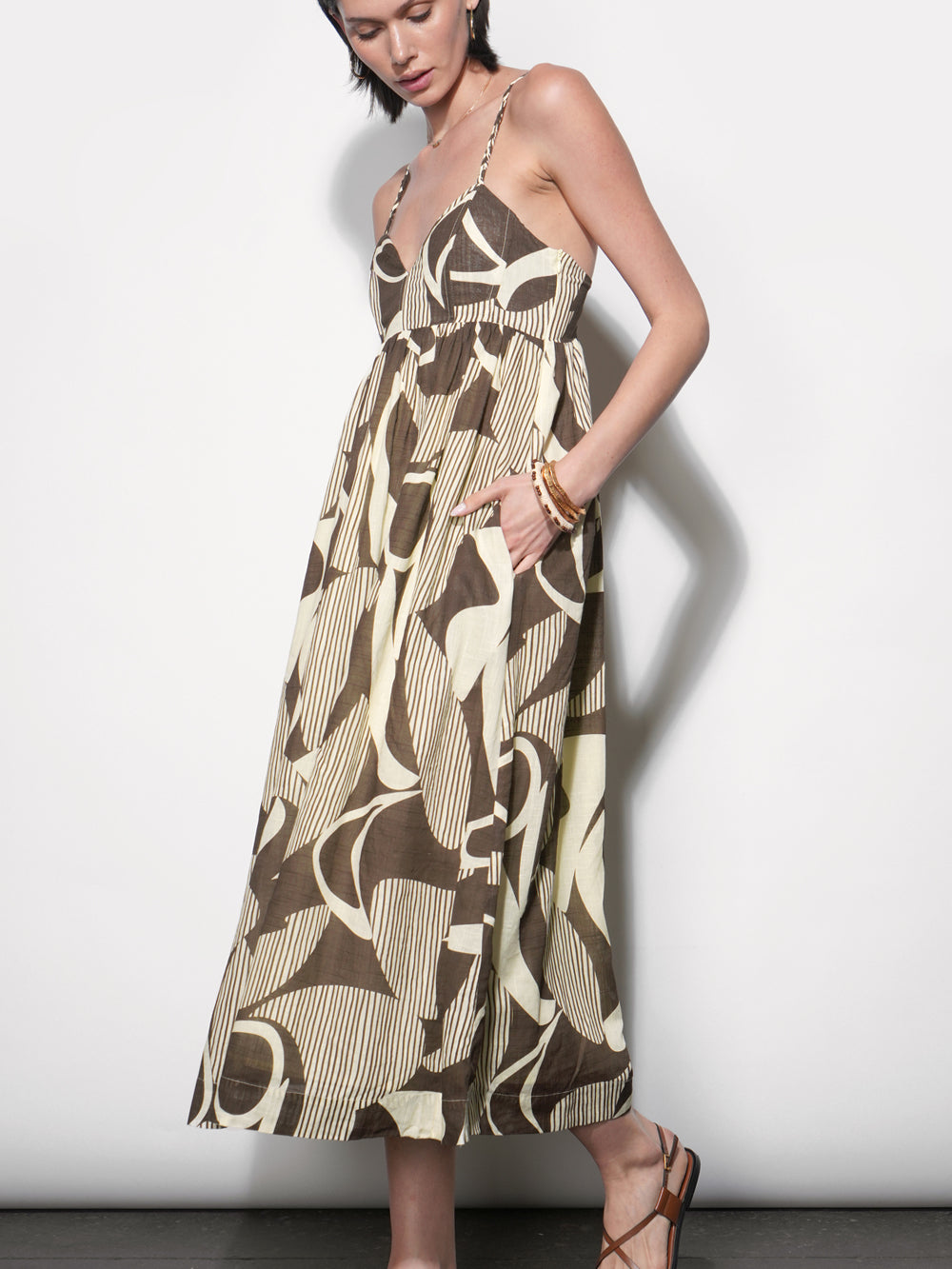 Midi dress in bold brown and cream abstract print with fine stripe texture, empire waist, thin straps, full gathered skirt, worn with tan strappy sandals and gold bangles, side