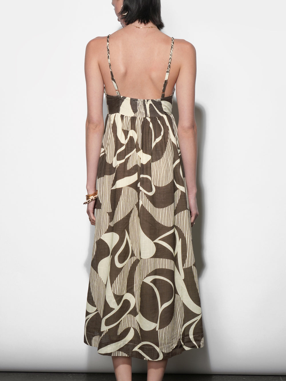 Back view of olive brown and cream abstract-print midi dress with open back, wide smocked waistband, thin woven straps, full gathered skirt, worn with tan leather sandals.
