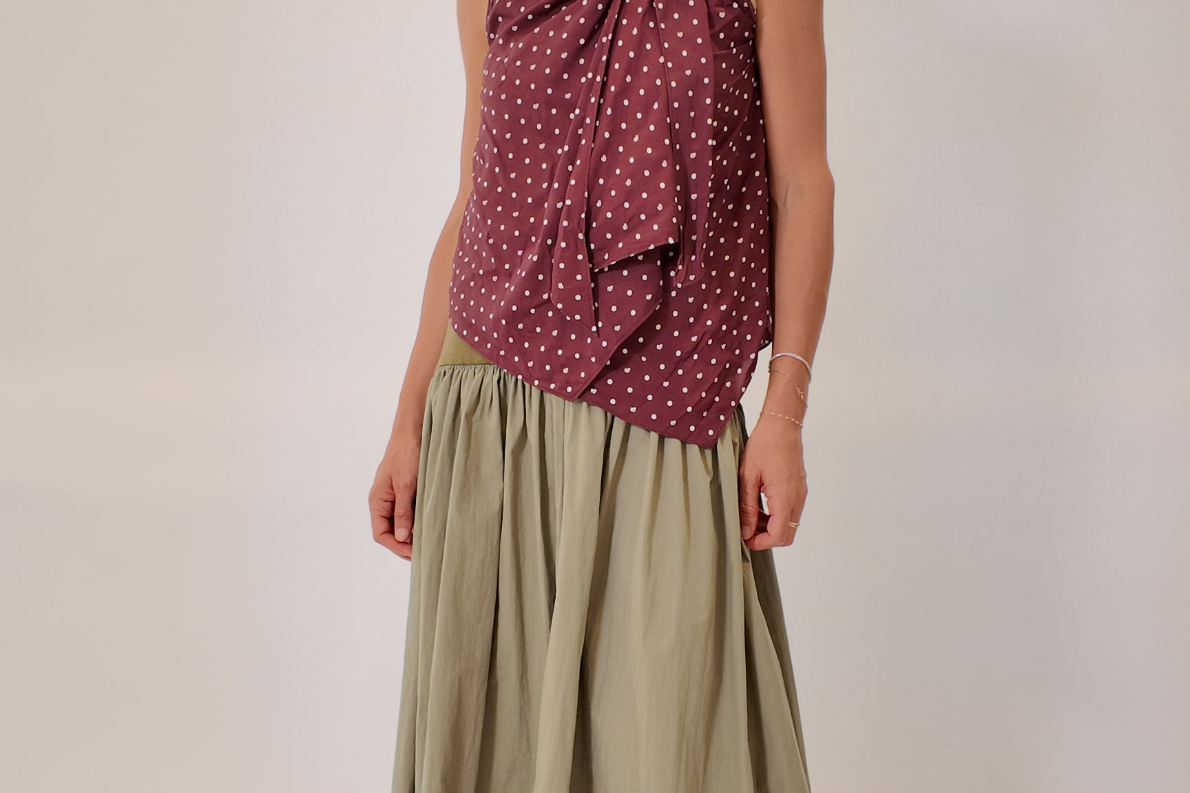 Sage green gathered maxi skirt styled with a burgundy polka dot sleeveless top and tan leather slide sandals, full-length silhouette with soft volume, front-facing model view.