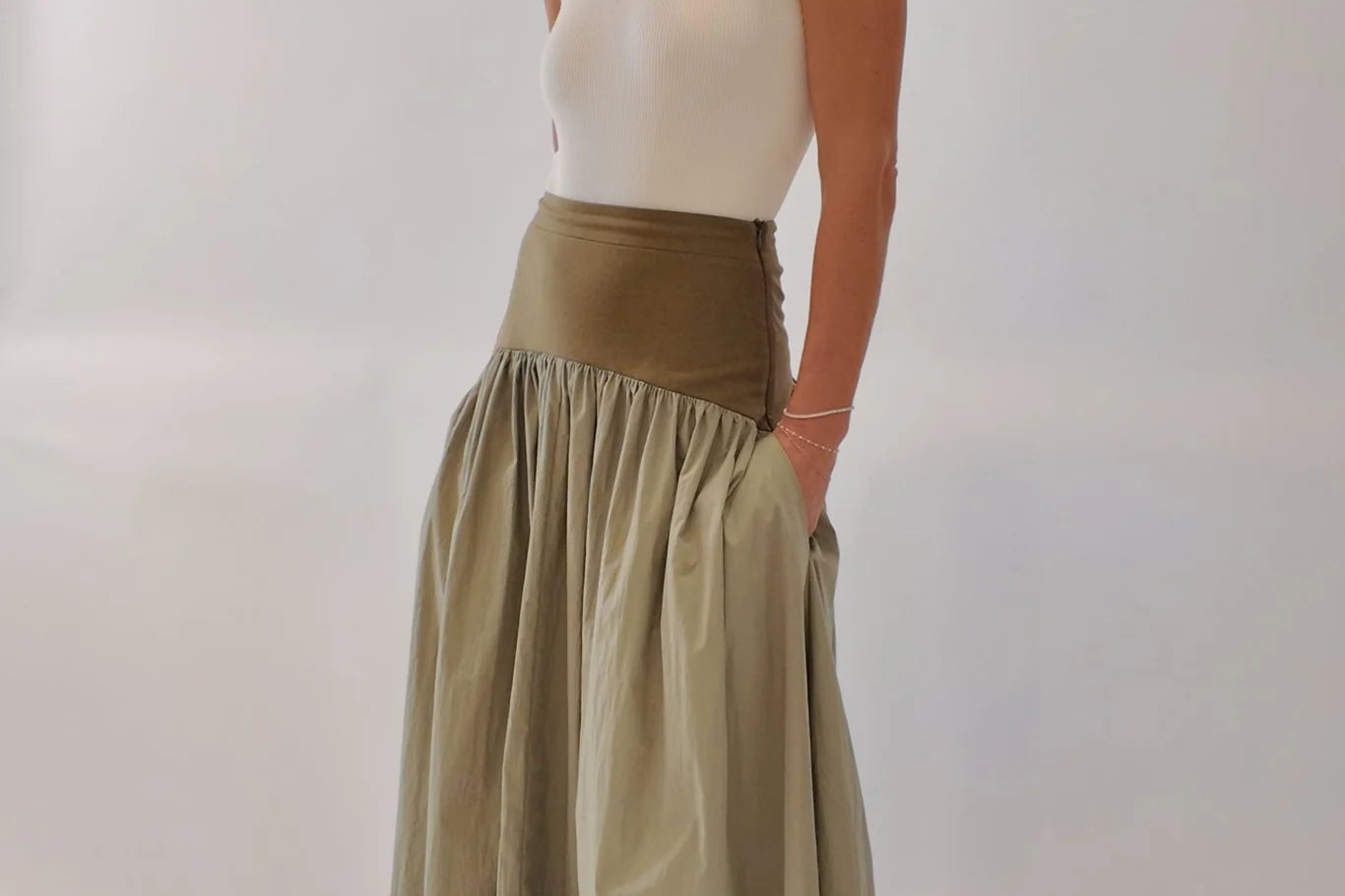 Sage green maxi skirt with structured olive waistband and voluminous gathered skirt, styled with cream ribbed tank and tan block-heel mules, three-quarter view showing side pocket