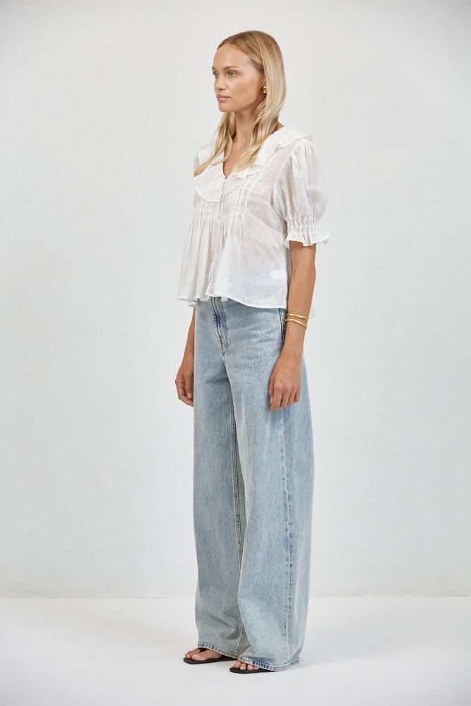 Side view of flowy white cotton-linen top with ruffle collar and smocked cuffs, tucked into light-wash wide-leg jeans, gold b