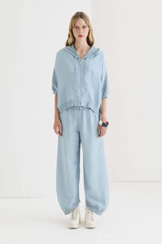 Relaxed slate blue cotton jogger trouser with gathered cuffed hem, elastic drawstring waist, worn with matching zip hoodie and white sneakers, full-length front view.