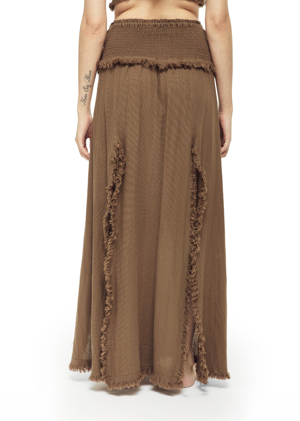 Back view of a warm brown cotton maxi skirt with smocked elastic waistband, raw fringe trim along vertical panel seams, and frayed hem. Loose A-line drape, open-weave texture