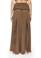 Back view of a warm brown cotton maxi skirt with smocked elastic waistband, raw fringe trim along vertical panel seams, and frayed hem. Loose A-line drape, open-weave texture