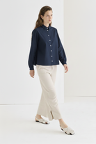 Navy cropped boxy cotton poplin shirt with pearl buttons, worn untucked over cream straight-leg trousers, styled with woven white mule flats, three-quarter walking view.