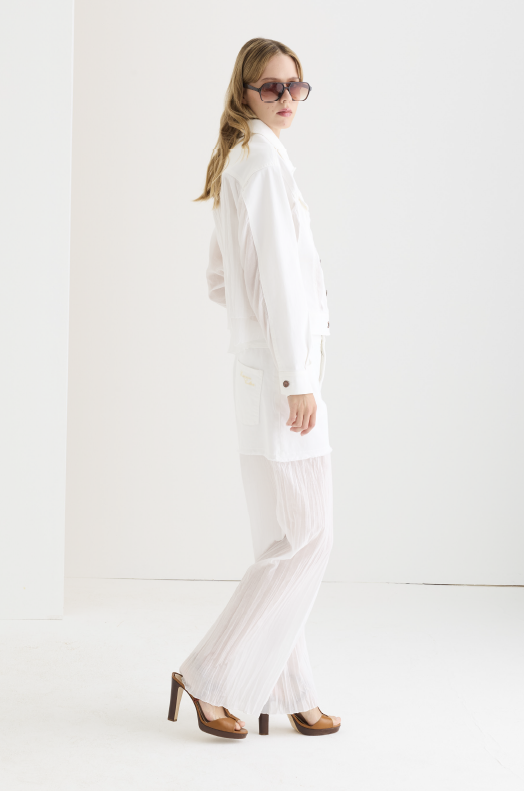 White structured coat with tailored lapel, patch pockets, and tortoiseshell buttons, styled over sheer white wide-leg trousers with brown platform heels and oversized sunglasses.