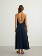 Navy halter maxi dress, back view — deep open V with double low strap, matte button at neck, A-line silhouette, textured slub fabric, black strappy sandals.