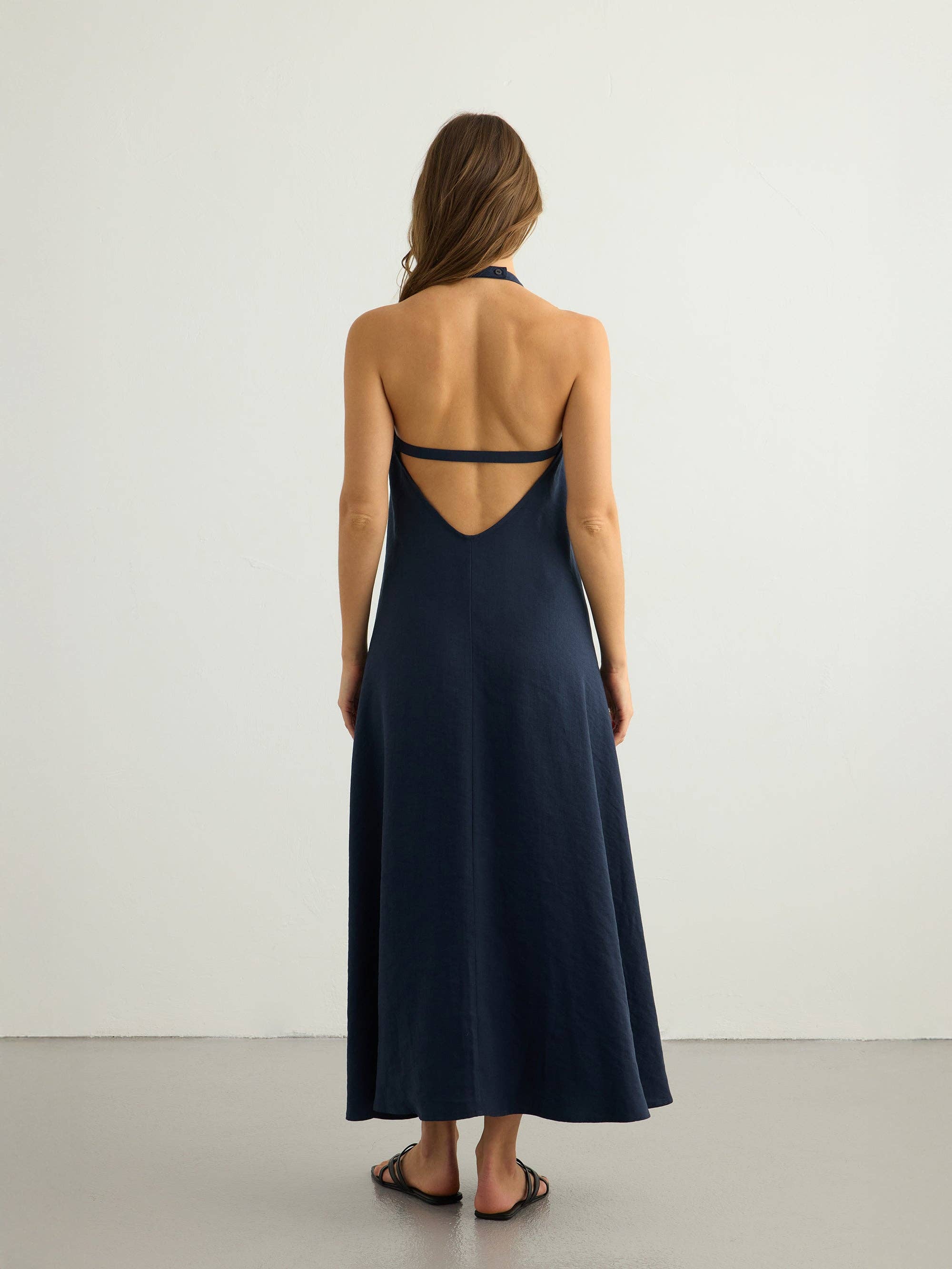 Navy halter maxi dress, back view — deep open V with double low strap, matte button at neck, A-line silhouette, textured slub fabric, black strappy sandals.