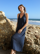 Navy slub halter maxi dress, deep V-neckline, relaxed A-line fit, worn against sea rocks on a California beach, bright blue sky and ocean behind.