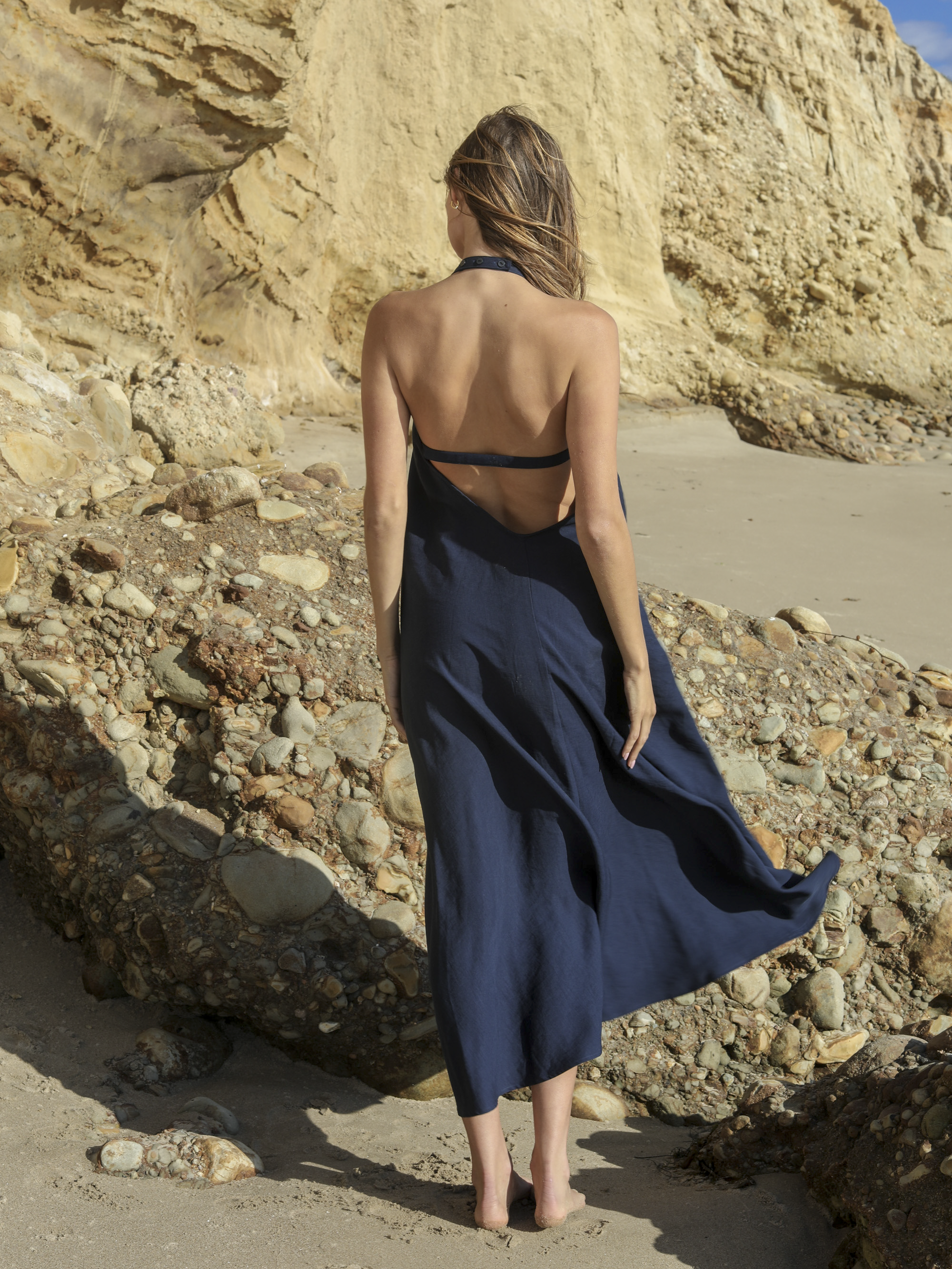 Navy linen halter maxi dress, deep open back with double strap and matte button closure, flowing A-line skirt caught in ocean breeze, photographed from behind on rocky California beach