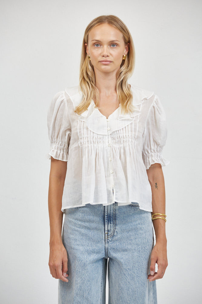 White sheer cotton-linen blouse with ruffle collar, pin-tuck bib, smocked puff sleeves, styled with wide-leg light-wash jeans