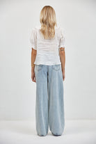 Back view of sheer white cotton-linen top with smocked puff sleeves and ruffled hem, tucked into light-wash wide-leg jeans.