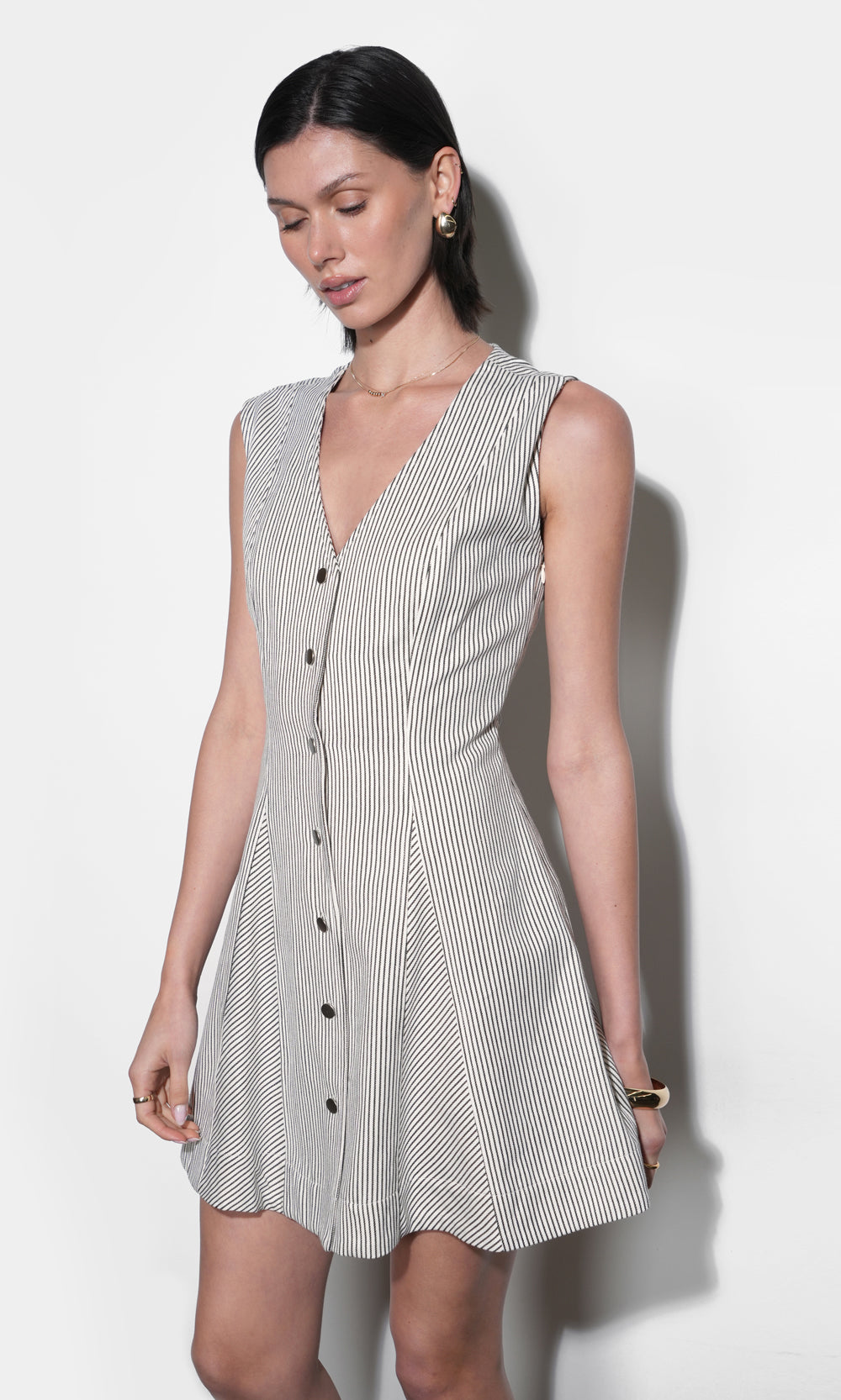 Sleeveless cream and black pinstripe mini dress with deep V-neckline, matte brass snap buttons, and angled panel skirt — fitted waist, front view, mid-thigh length.