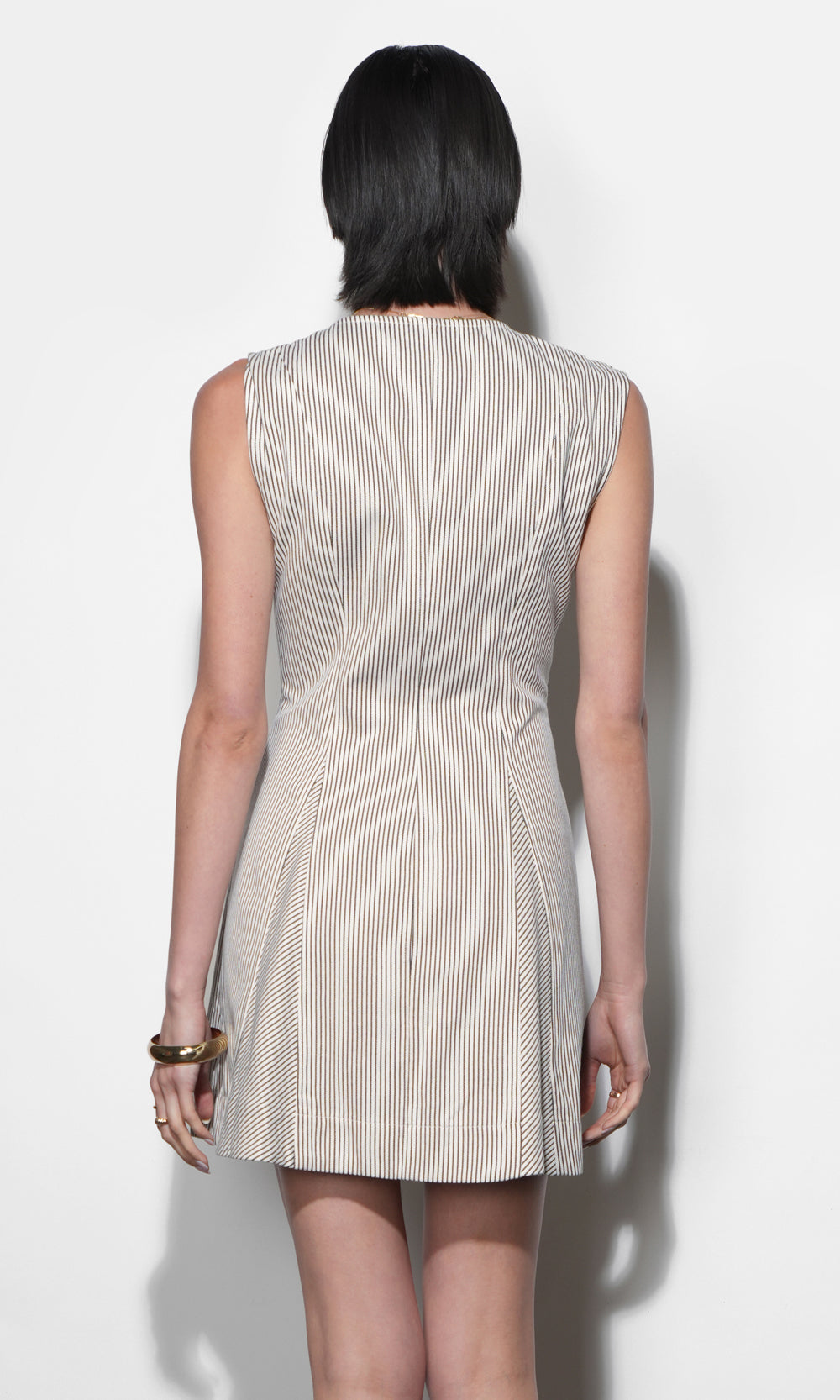 Back view of cream and near-black ticking stripe mini dress, sleeveless, structured canvas fabric, angled skirt panels, fitted waist, gold bangle bracelet detail.