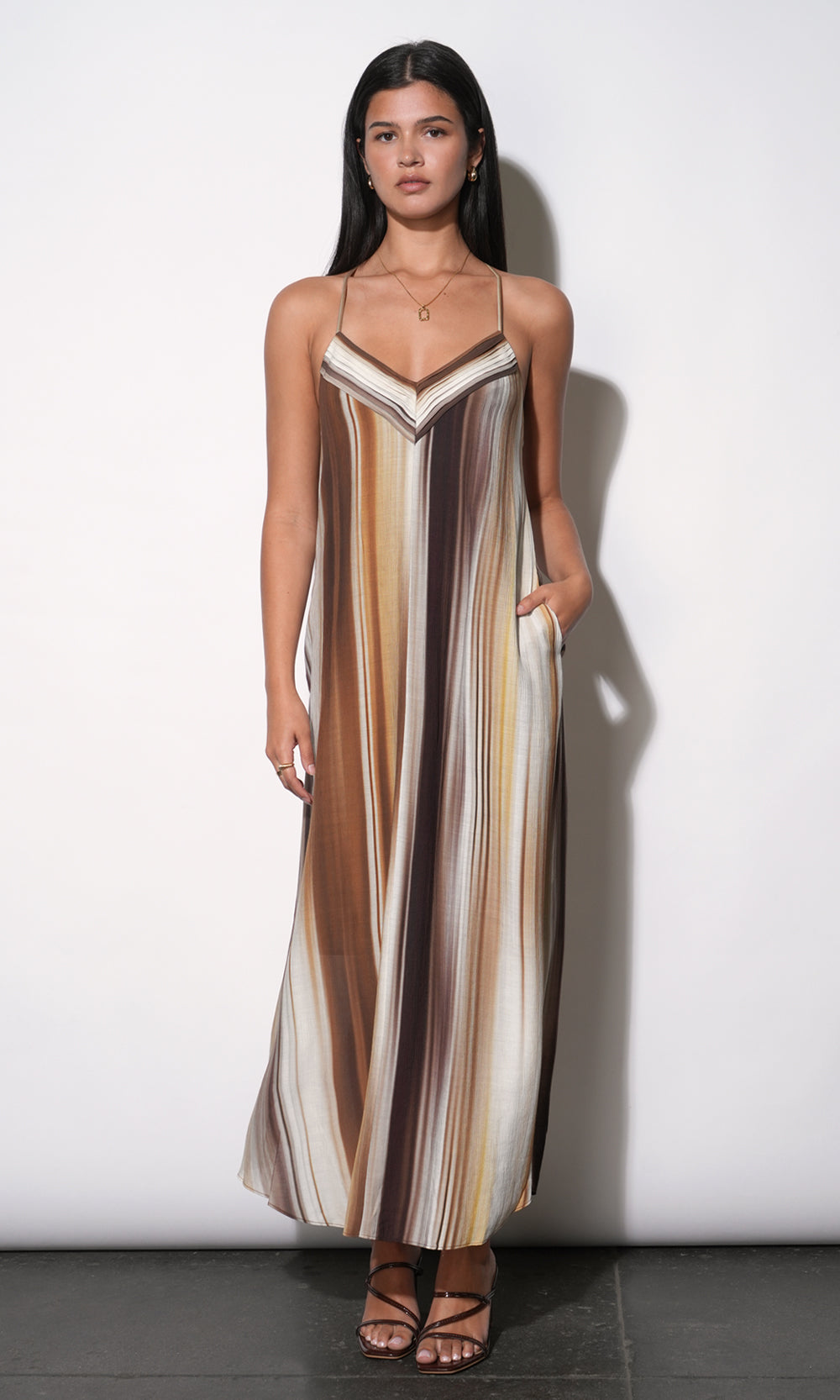 Fluid maxi dress with vertical watercolor stripe print in warm earth tones, V-neckline with contrast binding, thin spaghetti straps, relaxed A-line silhouette to floor, side pocket visible, paired