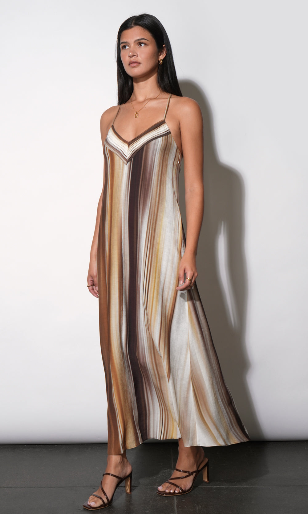 Fluid satin-finish maxi slip dress with blended vertical watercolor stripe print, V-neckline with contrast binding, spaghetti straps, full-length A-line skirt. Side profile. Styled with strappy
