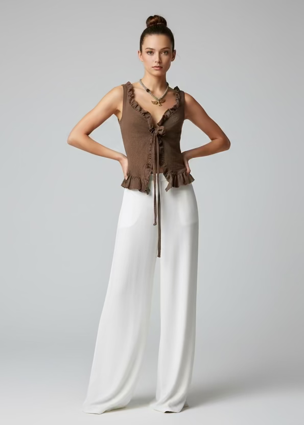 Warm walnut brown smocked ruffle top with tie-front V-neck, worn with wide-leg white trousers, hair up, gold layered necklace