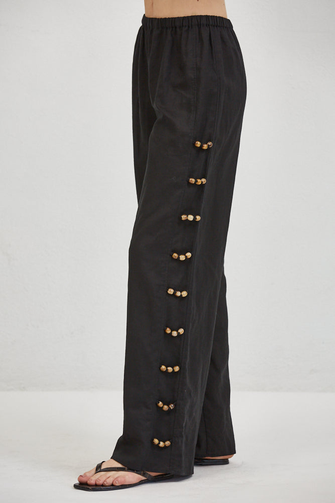 Linen wide-leg trousers, side view showing full-length row of paired gold knotted buttons along outer seam