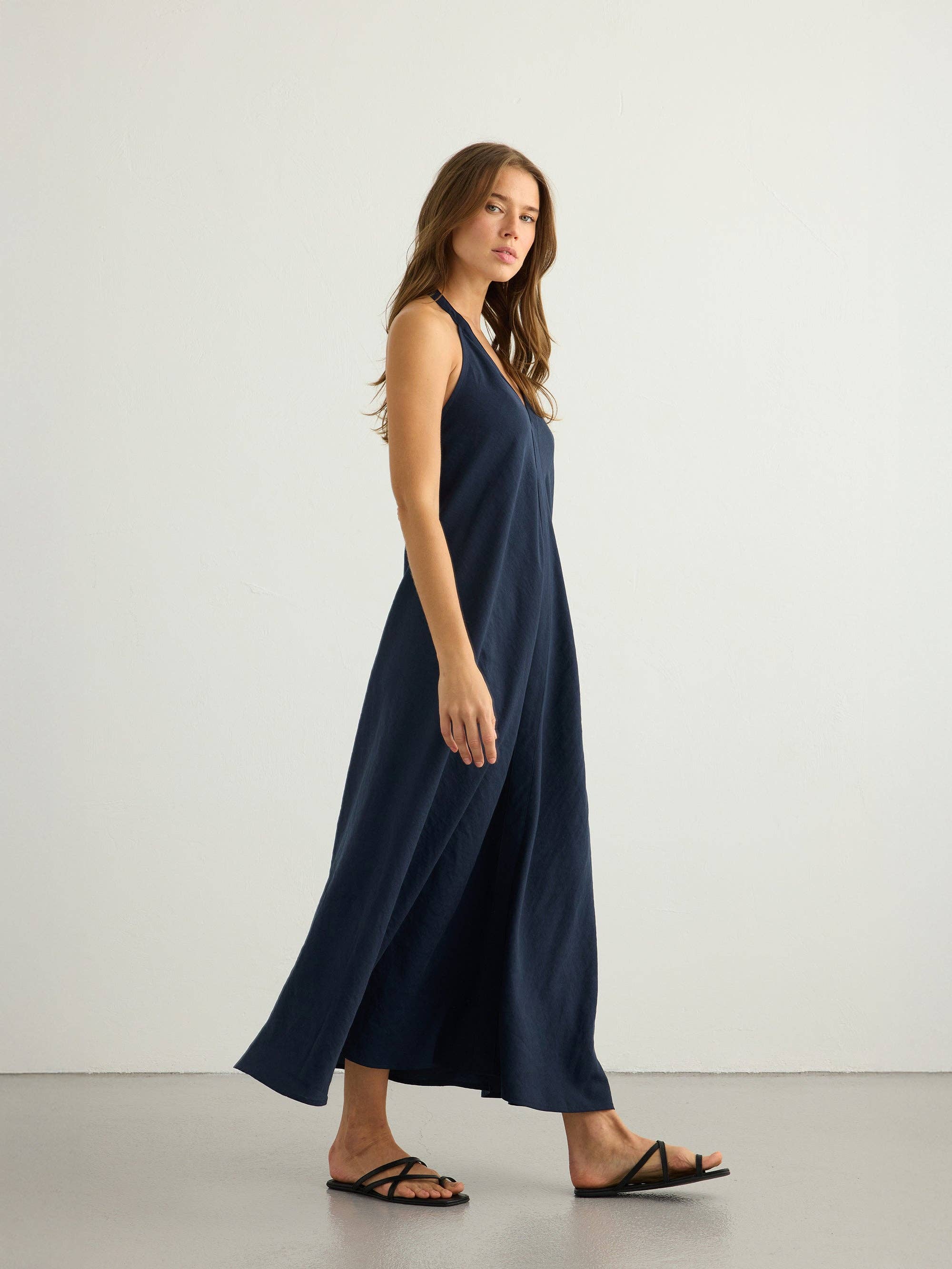 Navy halter maxi dress, side profile, fluid A-line silhouette, floor-length hem, natural slub fabric, black strappy flat sandals, clean center front seam