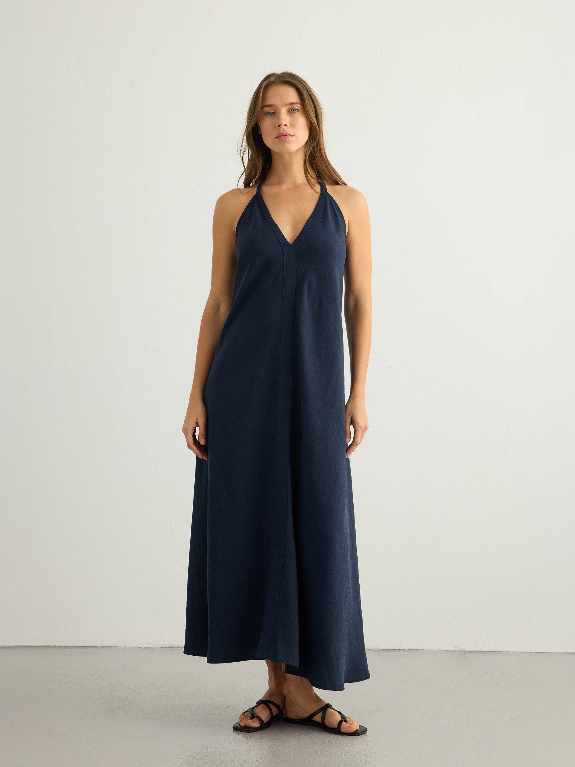 Navy halter maxi dress, deep V-neck, clean center front seam, A-line silhouette in textured slub fabric, paired with black strappy flat sandals.