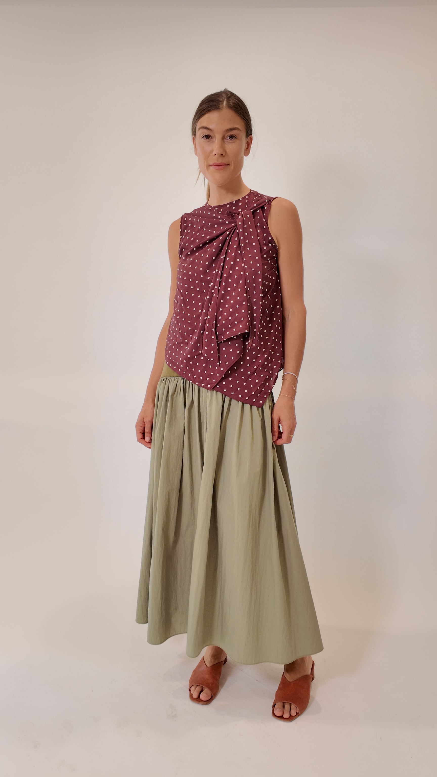 Sage green gathered maxi skirt styled with a burgundy polka dot sleeveless top and tan leather slide sandals, full-length silhouette with soft volume, front-facing model view.