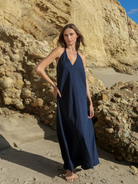 Navy halter V-neck maxi dress in textured slub fabric, floor-length A-line silhouette, worn barefoot on sandy beach against sandstone cliffs.