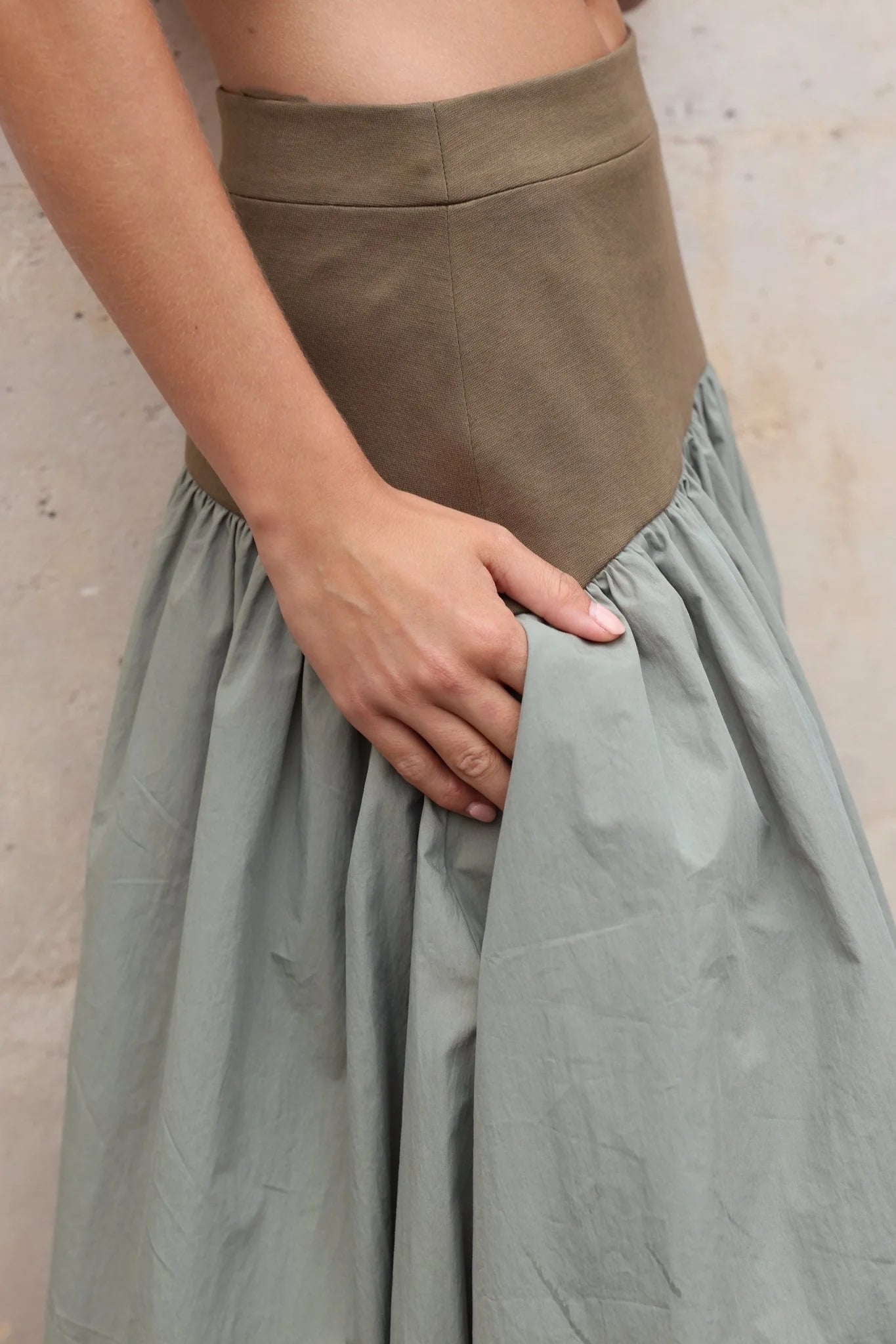 Close-up of a deep side pocket on a sage green gathered cotton skirt, hand pulling fabric open to reveal pocket depth, khaki structured waistband above.