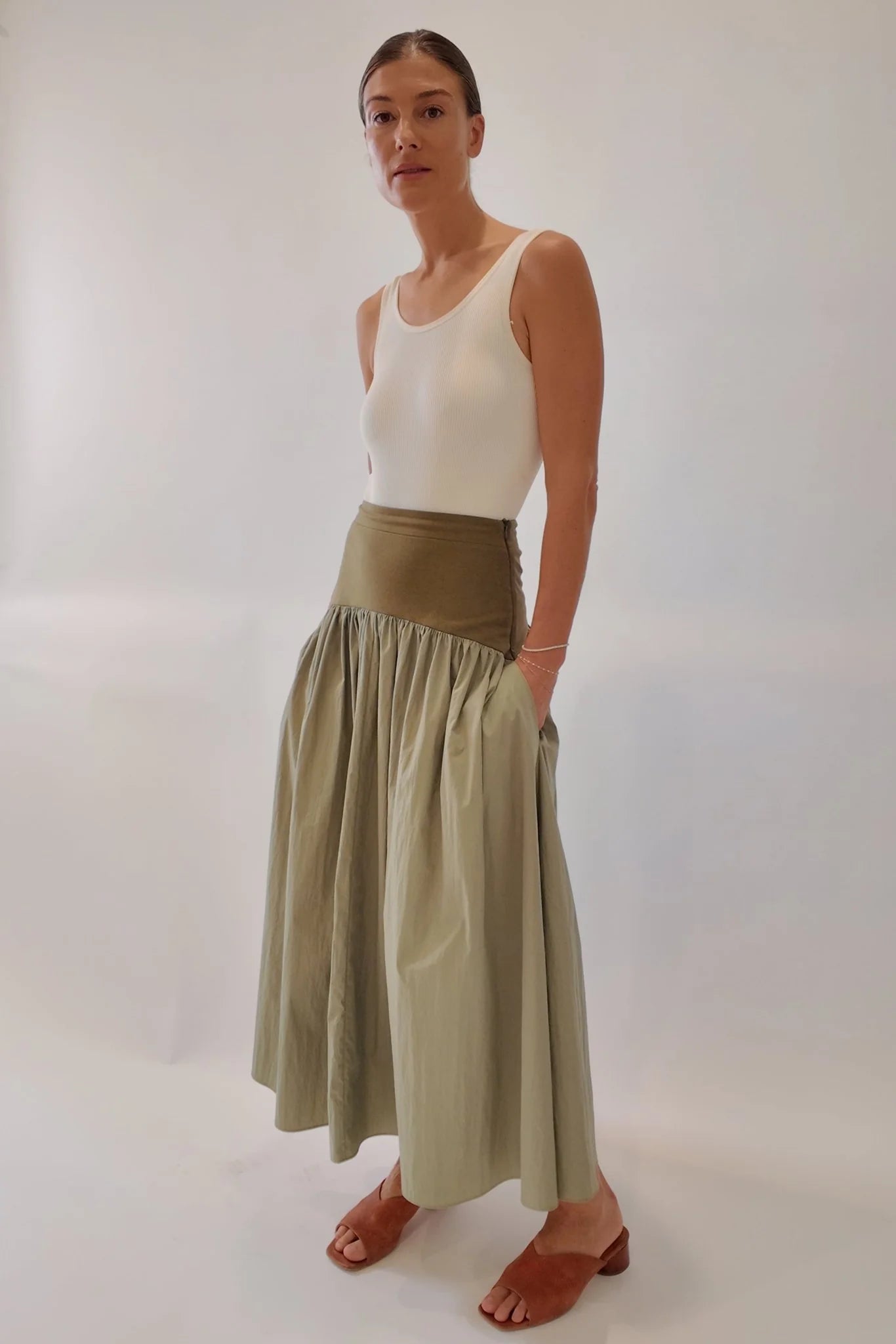 Sage green maxi skirt with structured olive waistband and voluminous gathered skirt, styled with cream ribbed tank and tan block-heel mules, three-quarter view showing side pocket