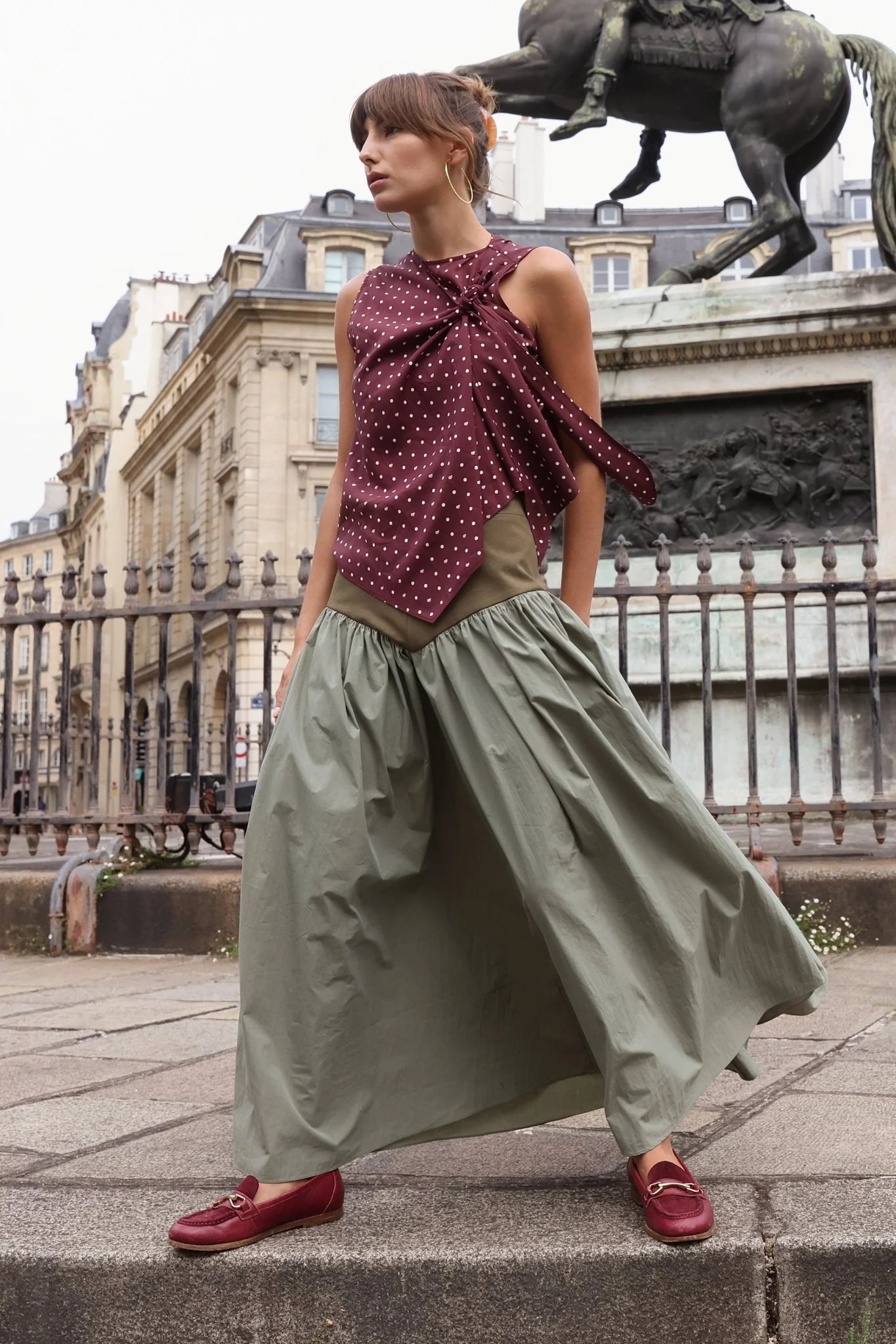 Sage green full-length gathered maxi skirt with olive fitted waistband, styled with burgundy polka dot bow top and burgundy loafers, shot outdoors in Paris near an equestrian